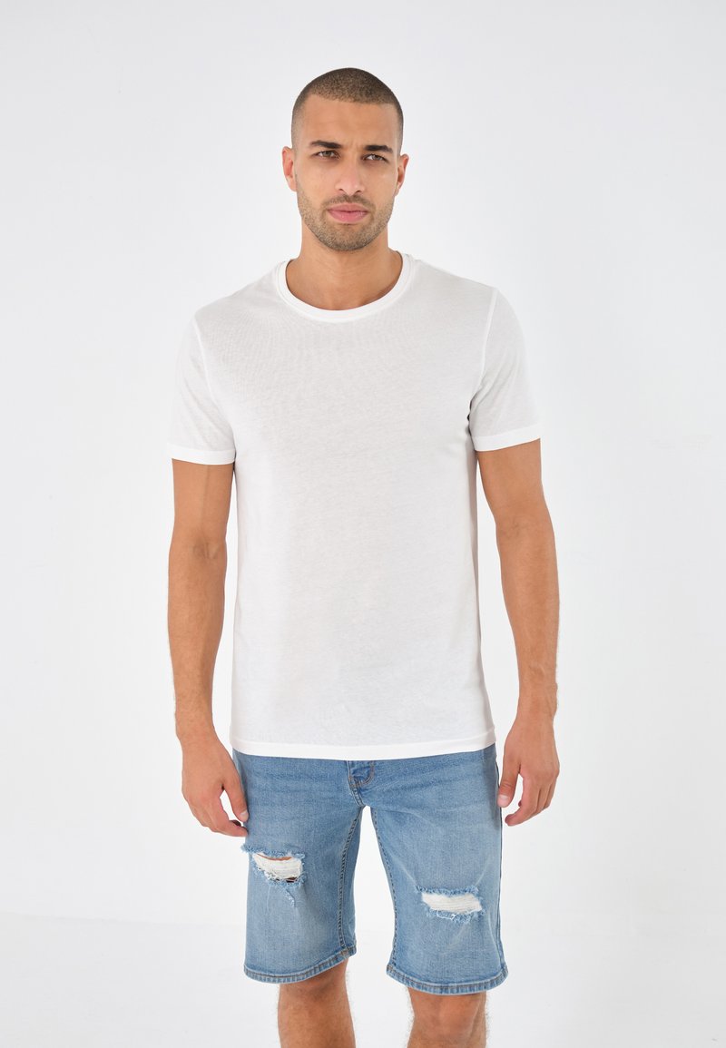 T-shirt bianca a maniche corte con scollatura rotonda e rifiniture a contrasto, abbinata a pantaloncini di jeans azzurri chiari con dettagli sfilacciati.