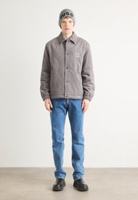 Jachetă de corduroy gri cu logo-ul HUGO, blugi albaștri și căciulă tricotată gri, având o croială lejeră și închidere cu buton. Pantofi negri completează ținuta.