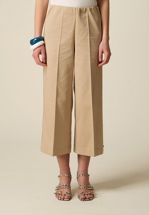 Beige vidde-bukser i cropped model båret med slangeprintede remsandaler og chunky blå og hvide armbånd på venstre håndled.