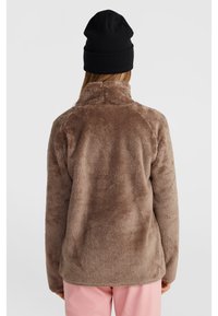 Fleece jack in zacht bruin met een hoge kraag en lange mouwen, gecombineerd met lichtroze broek. Een zwarte beanie maakt de look compleet.