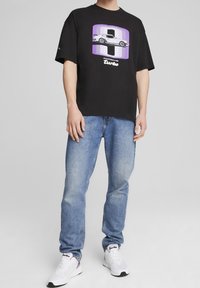 Camiseta negra de manga corta con un gráfico de Porsche en púrpura y blanco, combinada con jeans de tela azul claro y zapatillas blancas con acentos en púrpura.