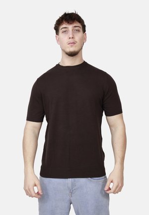 Giovane con capelli corti scuri, indossa una maglietta nera a maniche corte e jeans azzurri, in piedi davanti a uno sfondo bianco.