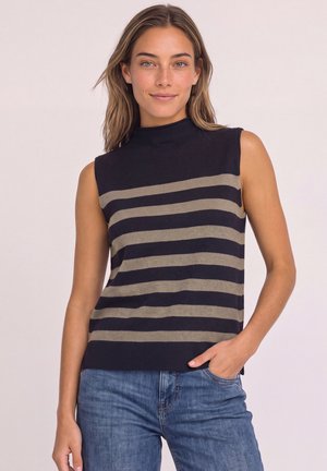 Femme portant un haut noir sans manches avec des rayures beiges horizontales et un jean bleu, debout avec une main dans la poche sur un fond uni.