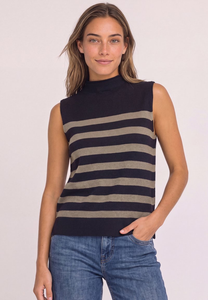 Femme portant un haut noir sans manches avec des rayures beiges horizontales et un jean bleu, debout avec une main dans la poche sur un fond uni.