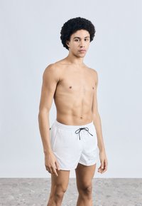 Valged ujumispüksid elastse vöökohaga, musta paelaga ja esiküljel reljeefne logo. Sile tekstuur, keskmise pikkusega reieni.