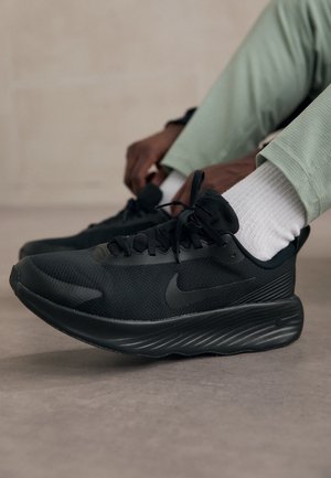 Osoba wiążąca czarne buty do biegania Nike w białych skarpetkach i jasnych zielonych spodniach na gładkiej podłodze wewnątrz pomieszczenia.