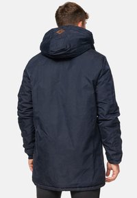 Parka bleu marine avec une grande capuche, tissu texturé et patch en cuir au dos. Manches longues et coupe droite.