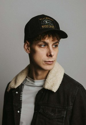 Jauns vīrietis ar cirtainiem matiem, valkājot melnu cepuri ar uzrakstu "BLACKSKIES RONIN DUST" un melnu jaku ar bēša krāsas kažokādas apkakli, skatās uz sāniem.