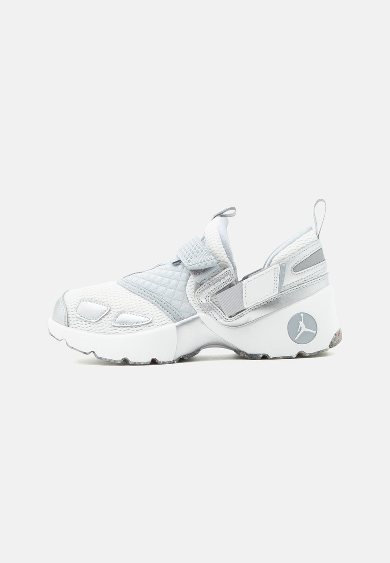 Zapatilla deportiva blanca y gris sin cordones, con malla, tejido acolchado, correas ajustables y un logotipo circular en el talón.