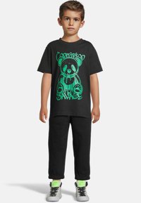 Juodas marškinėliai su žaliu meškino grafiniu atvaizdu ir tekstu, derinami su juodomis kelnėmis ir pilkais sportbačiais su neoninėmis žalios spalvos akcentais. Kasdienis stilius ir laisvas siluetas.