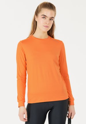 Ung kvinde med langt hår iført en orange langærmet trøje og sorte cykelbukser, stående mod en ensfarvet hvid baggrund.