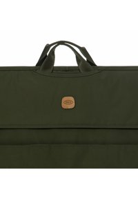 Bolsa de tela verde oliva con asas superiores dobles, que presenta un acento de logo de cuero marrón y un bolsillo con cremallera horizontal en la parte delantera.