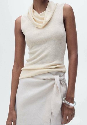 Mouwloos crème top met gedrapeerde kraag, ingestopt in een lichtbeige rok met strik, gecombineerd met een dikke transparante armband aan de rechterpols.