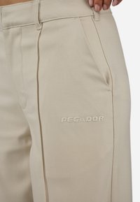 Beige pants made of textured fabric with front pleats. Features an embroidered "PEGADOR" logo on the left side.