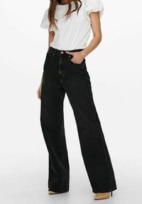 Jeans anchos negros de mezclilla, de cintura alta, con un corte relajado. Combinados con una blusa blanca de manga corta y tacones beige.