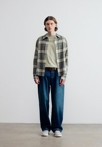 Νέος άνθρωπος όρθιος, φορώντας μπεζ μπλούζα με σχέδιο, γκρι και μαύρο πουκάμισο καρό, φαρδιά μπλε τζιν, λευκά αθλητικά παπούτσια, πάνω σε απλό λευκό φόντο.