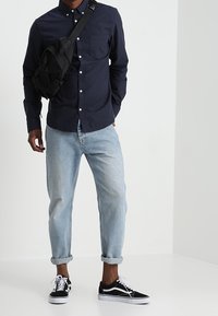 Granatowa koszula na guziki, jasnoniebieskie dżinsy z podwiniętymi nogawkami, czarno-białe trampki oraz czarna torba na ramię. Prosty design z funkcjonalnymi detalami.