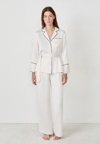 Jedwabny komplet piżamowy w miękkim odcieniu kremowym z czarnym wykończeniem. W skład zestawu wchodzi zapinana na guziki koszula z kieszonką na piersi oraz szerokie spodnie związywane w talii.