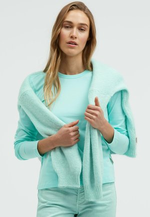 Frau trägt hellblaues langärmliges Shirt und Hose, hält einen passenden weichen Strickpullover, der über ihre Schultern gelegt ist.