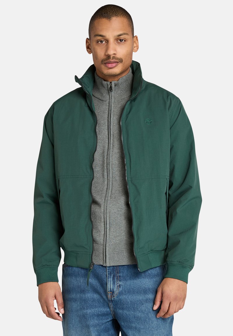 Grüne, leichte Jacke mit Stehkragen, Reißverschluss vorne und elastischen Bündchen, übereinander getragen mit einem grauen Kapuzenpullover mit Reißverschluss.