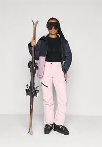 Skiausrüstung: schwarze und pinke Jacke, schwarzes Oberteil, pinke Skihosen, schwarze Skischuhe; hält Holzski mit Bindungen, schwarze Skibrille und geflochtenes Haar.