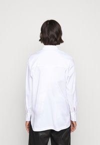 Camicia bianca a maniche lunghe realizzata in tessuto morbido, con colletto alto, polsini con bottone e fessure laterali all'orlo.