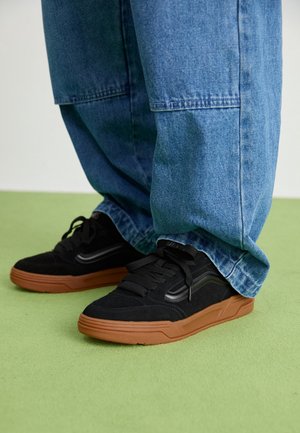 Zapatillas negras con suelas marrones usadas con vaqueros azul claros y sueltos, de pie sobre un suelo verde claro.