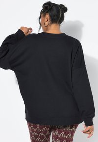 Sudadera negra de gran tamaño con mangas largas, puños de canalé y escote redondo. Combinada con leggings ajustados y estampados. Vista trasera sencilla.
