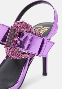Sandale à talon haut violette brillante avec boucle métallique ornée gravée "Versace Jeans Couture" sur la semelle intérieure noire.