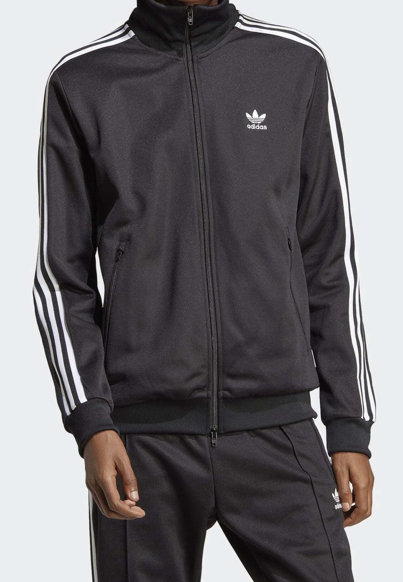 Person som bär en svart Adidas träningsoverall med vita ränder på ärmarna och byxorna, jacka med dragkedja, och Adidas-logotyp på bröstet och låret.