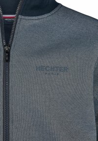 Close-up van een navy en witte textuur zip-up jack met "HECHTER PARIS" gedrukt op de borst en een bijpassende ritssluiting.