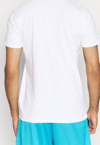 Hombre que lleva una camiseta blanca de manga corta lisa y unos pantalones cortos de malla azules brillantes, visto desde atrás contra un fondo liso.