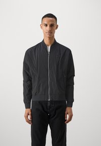 Veste bomber noire en matériau léger, avec un zip sur le devant, des poignets côtelés, des poches latérales et un patch logo sur la poitrine.