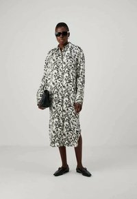 Pessoa vestindo um vestido midi floral preto e branco, sapatos loafers pretos, óculos de sol e segurando uma mala preta em fundo plain.