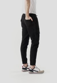 Pantalones cargo negros con puños elásticos, múltiples bolsillos y corte ajustado, combinados con zapatillas grises y blancas con detalles en rojo.