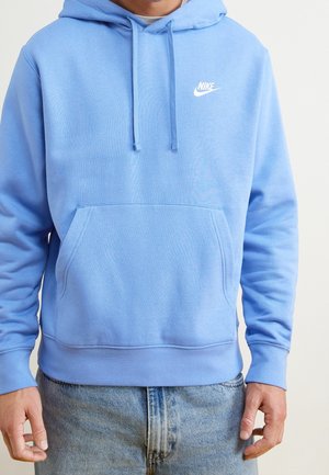 Mężczyzna w jasno-niebieskiej bluzie Nike z kapturem, kieszenią z przodu i sznurkami, ubrany w jasno-niebieskie dżinsy, na jednolitym tle.