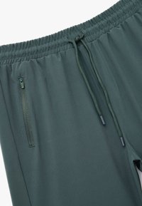 Pantalones deportivos de color verde oscuro con una cintura elástica, cordón de ajuste y bolsillos con cremallera seguros, que cuentan con una textura de tela suave y ligera.