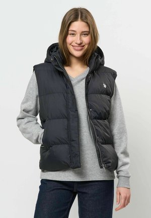 Jeune femme portant une veste matelassée noire à capuche par-dessus un pull gris et un jean foncé, souriant avec une main dans sa poche.