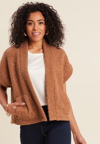 Brauner, strukturierter Cardigan mit kurzen Ärmeln und zwei vorderen Taschen, getragen über einem hellen, glänzenden Oberteil. Weiches Material mit dezenten Schimmereffekten.