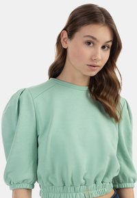 Világoszöld, cropped fazonú pulóver, puffos ujjal, rugalmas aljjal és puha tapintással. Kerek nyakkivágással és bordázott részletekkel rendelkezik.