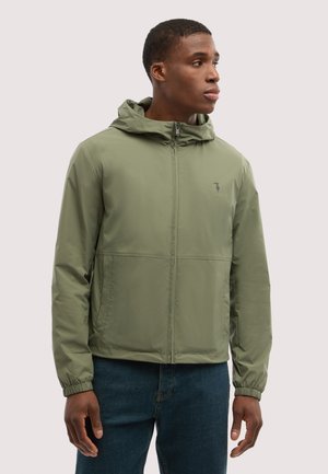 Mand iført olivengrøn hættetrøje med lynlås og mørkeblå jeans, der ser mod højre mod en ensfarvet lys baggrund.