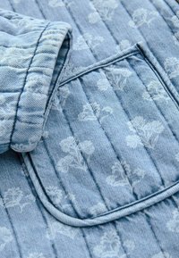 Sininen denim-kangas, jossa on valkoinen kukkakuvio, varustettuna vuoratulla taskulla ja teksturoidulla ompelulla. Lähikuva kauluksesta ja taskun yksityiskohdista.
