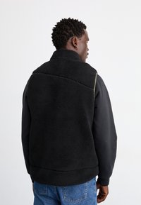 Gilet en polaire noire avec un col haut, surface texturée et coupe décontractée, porté par-dessus une chemise noire à manches longues et un jean bleu.