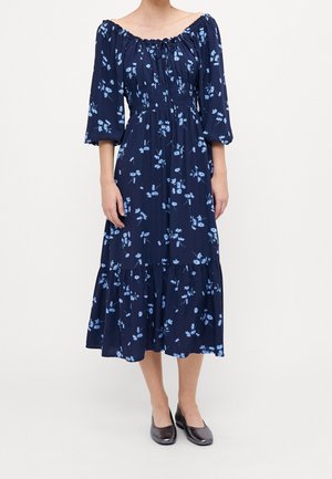 Kvinde iført en navyblå off-shoulder midi-kjole med lyseblåt blomsterprint, trekvartærmer og sorte flade sko.