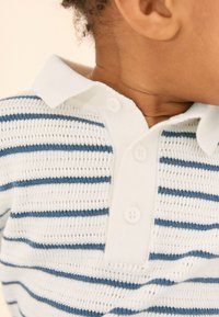 Randig pikétröja med vita och blå horisontella ränder, knäppt krage, tillverkad av stickat tyg med en texturerad yta.