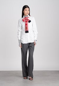 Γυναίκα με λευκή πουκαμίσα με πτυχώσεις, γκρι παντελόνι, ροζ μαντίλι με αρκούδα και καρδιές, γκρίζια γάντια χωρίς δάχτυλα και μαύρες ανοιχτές μπότες, που στέκεται σε γκρι πάτωμα.