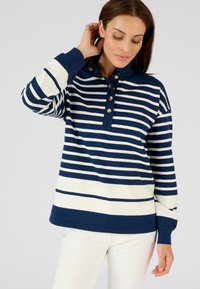 Polo a maniche lunghe a righe blu navy e bianche con colletto, tre bottoni in rame e polsini a costine. Il tessuto sembra morbido e dalla texture piacevole.