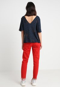 Navy-Bluse mit kurzen Ärmeln und V-Ausschnitt, ausgestattet mit einem goldfarbenen Punktmuster. Kombiniert mit roten Slim-Fit-Hosen und silbernen Sneakern.