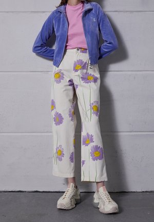 Personne portant un pantalon crème à imprimé floral violet et jaune, une veste zippée lavande, un haut rose et des baskets blanches épaisses devant un mur gris.