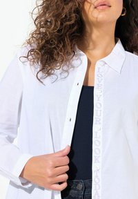Witte blouse met geborduurde tekst "LOVE YOUR LOOK TODAY" op de knoopsluiting, gedragen over een marineblauwe top, met een gladde, lichte stof.